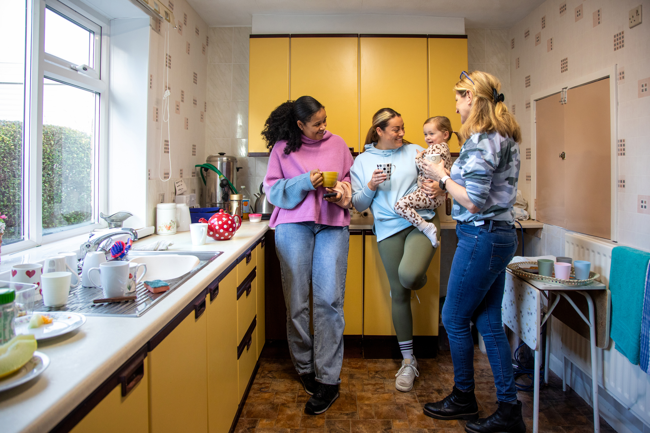 Full shot of a group of adults standing in a community kitchen smiling and talking to each other, The woman in the middle is holding her baby in her arms. The institute is located in Seghill, Northumberland.